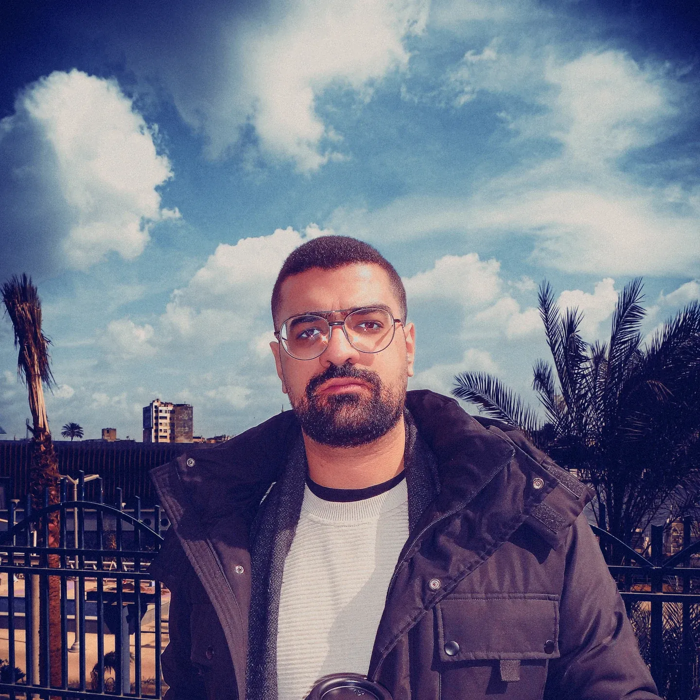 Bassem wearing glasses and a dark jacket outdoors beneath a cloudy sky